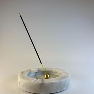 May include: A white marble incense holder with a gold-coloured ball and a black incense stick. The holder is round with a recessed area for catching ash. The incense stick is angled upwards, creating a minimalist aesthetic.