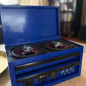 May include: A custom-built, blue speaker box with two speakers visible. The box has a hinged lid and a drawer-like front with black handles. It features USB ports and control switches, and is designed for audio playback.