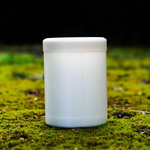 以下が含まれることがあります： 白い円筒形の容器と、それに合う蓋。容器は緑色の苔と暗い背景を背景に配置されています。容器はプラスチック製のように見えます。
