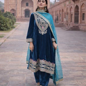 May include: A woman in a dark blue velvet salwar kameez, featuring detailed embroidery on the neckline, sleeves, and hem. A light blue dupatta with gold trim is draped over the shoulders. The outfit is worn with matching trousers.