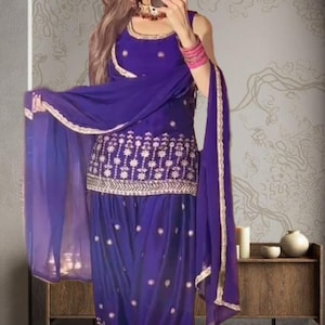 May include: A vibrant purple salwar kameez ensemble. The outfit features intricate gold embroidery on the top and bottom, with a matching sheer dupatta. The outfit is a traditional South Asian style.