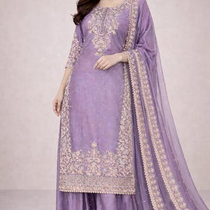 Könnte beinhalten: Ein lavendelfarbenes Salwar Kameez-Ensemble mit aufwendiger goldener Stickerei. Das Outfit umfasst eine lange Tunika, eine Hose mit weitem Bein und einen passenden, durchsichtigen Dupatta. Die Stickerei konzentriert sich auf Ausschnitt, Saum und Dupatta-Ränder.