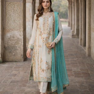 Puede incluir: Un salwar kameez bordado de color marfil con un dupatta a juego. La túnica y los pantalones de manga larga están adornados con bordados florales en colores pastel. Un dupatta turquesa con ribete dorado añade un toque de color. El conjunto se lleva con tacones a juego.
