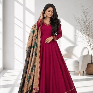 Puede incluir: Un vestido largo y fluido de color burdeos con ribetes dorados y mangas largas. Una chal con estampado de diseños florales verdes y negros se coloca sobre el vestido. El escote y las mangas tienen bordados intrincados.