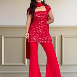 Könnte beinhalten: Ein leuchtend rotes Ensemble, bestehend aus einem quadratischen Oberteil mit goldenem Muster, ausgestellten Hosen und einem Schal. Das Outfit ist in einem kräftigen, einfarbigen Rot gehalten und wirkt dadurch harmonisch. Das Oberteil hat kurze Ärmel, die Hose einen weiten Beinverlauf.