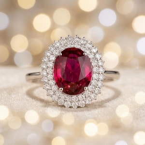 Puede incluir: Un anillo con una gran piedra preciosa de color rojo intenso y corte ovalado en el centro, rodeada por un doble halo de pequeñas piedras transparentes de corte redondo. La montura es una banda de metal plateado pulido.