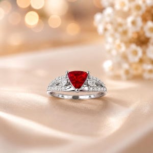 May include: A silver ring featuring a striking, triangular, deep red gemstone. The band is embellished with small, clear accent stones. The ring is displayed against a soft, neutral backdrop, emphasizing its elegant design.