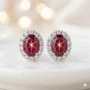 May include: A pair of oval-shaped ruby earrings with a halo of small, clear gemstones. The rubies have a deep red color and are surrounded by a ring of sparkling white stones. The earrings are set in a silver-colored metal.