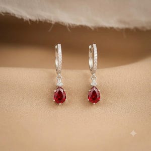 May include: A pair of silver hoop earrings featuring a row of small clear stones along the hoop and a dangling teardrop-shaped red gemstone pendant. Each pendant is accented with a tiny clear stone at the top where it connects to the hoop.