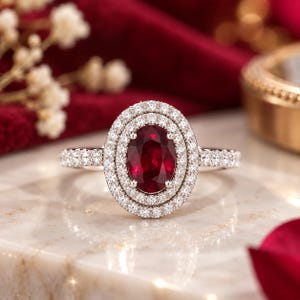 Op de afbeelding: Een ring met een centrale ovaal geslepen dieprode edelsteen, omringd door een dubbele halo van kleine witte steentjes. De ringband is aan de zijkanten ook bezet met kleine witte steentjes, wat zorgt voor een heldere en reflecterende uitstraling.