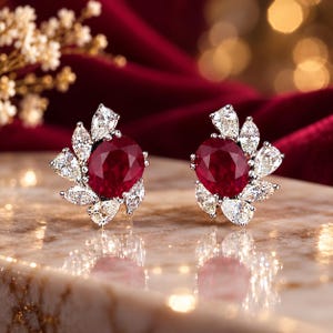 May include: A pair of stud earrings featuring a central oval-cut deep red ruby surrounded by a cluster of marquise and pear-cut clear white diamonds set in a metallic frame, creating a floral-inspired arrangement of gemstones.