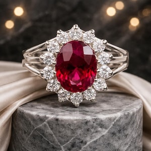 May include: A silver ring featuring a large, oval-cut ruby surrounded by a halo of smaller, round diamonds. The ruby is a deep red color, and the diamonds sparkle brightly. The ring is set on a gray marble surface.