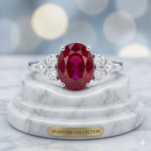 Könnte beinhalten: Ein silberner Ring mit einem großen, oval geschliffenen, tiefroten Rubin in der Mitte, flankiert von drei kleineren, rund geschliffenen weißen Diamanten auf jeder Seite. Der Ring steht auf einem weißen Marmorsockel mit einer goldenen Plakette GEMSTONE COLLECTION.