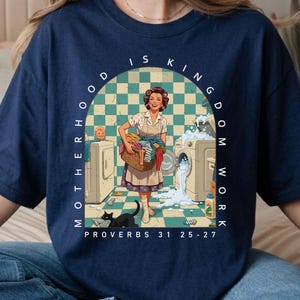 Puede incluir: Camiseta azul marino con una ilustración retro de una mujer lavando la ropa, un niño y un gato negro. El texto dice "Motherhood is Kingdom Work" y "Proverbios 31 25-27".