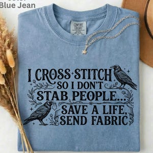 May include: Blue jean coloured t-shirt with the text "I CROSS STITCH SO I DON'T STAB PEOPLE... SAVE A LIFE, SEND FABRIC." The shirt features two crow illustrations and a star pendant necklace.