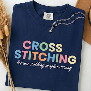 May include: A navy blue t-shirt featuring the words "CROSS STITCHING" in colourful letters. The phrase "because stabbing people is wrong" is written below in white script. A silver necklace with a star pendant is placed on the shirt.