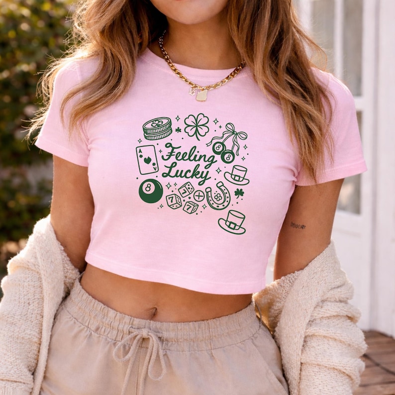 May include: A light pink crop top with a green graphic design featuring a four-leaf clover, dice, a horseshoe, and the words "Feeling Lucky". The shirt has short sleeves and a round neckline.