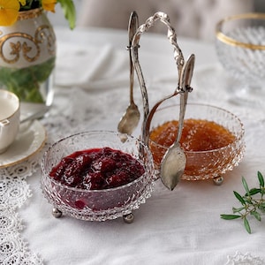 Op de afbeelding: Een decoratieve serveerset met twee glazen kommen gevuld met rode en oranje jam. De kommen staan in een zilverkleurige houder met twee lepels. De set staat op een wit kanten tafelkleed, met een vaas met bloemen op de achtergrond.