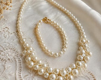 May include: A jewelry set consisting of a necklace and a matching bracelet, both featuring cream-colored round faux pearls of varying sizes. The necklace has a multi-layered cluster design at the front, and both pieces include gold-tone metal clasps.