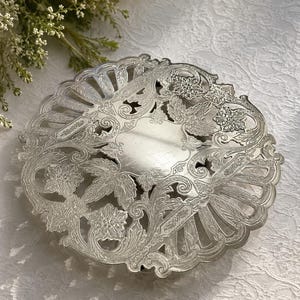Könnte beinhalten: Ein runder silberfarbener Metalluntersetzer mit einem komplizierten durchbrochenen Rand und einer glatten Mittelplatte. Die Oberfläche ist mit geprägten Blumenmotiven, geschwungenen Akanthusblättern und einem gewellten Rand mit strahlenförmigen Schlitzen verziert.