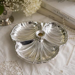 May include: A silver-toned metal serving dish shaped like a three-sectioned shell with a central candle holder. The dish features a scalloped edge and embossed radial lines radiating from the center, creating a distinct three-part compartment design.