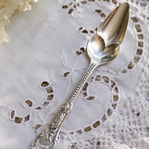 May include: A silver-toned metal spoon featuring a heart-shaped bowl and an elongated handle with intricate embossed floral and scrollwork detailing. The handle features a beaded border design that extends from the bowl to the decorative base.