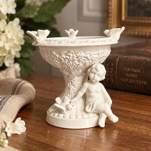 Peut inclure: Un bol sur pied en céramique blanche orné d'une figurine de chérubin sculptée assise à la base, tendant la main vers un petit oiseau. Le bol est décoré de grappes de raisin et de feuilles en relief, avec trois petits oiseaux sur le bord.