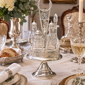 Puede incluir: Un soporte para condimentos plateado con un asa central y una base circular con grabados florales. El soporte sostiene cuatro recipientes de vidrio transparente, incluyendo dos saleros con tapas plateadas perforadas y dos vinagreras con tapones de vidrio.
