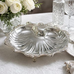 May include: A polished silver-plated serving dish shaped like a large scallop shell, featuring a scalloped edge with intricate embossed floral detailing and supported by three small, ornate feet. The surface is smooth and reflective.