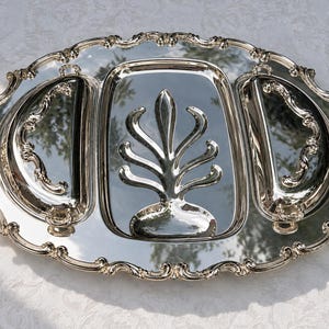 Puede incluir: Una bandeja de servicio ovalada de tono plateado con una superficie reflectante pulida. La bandeja presenta una sección central rectangular con un motivo vegetal estilizado en relieve y dos compartimentos semicirculares, bordeados por un borde festoneado.