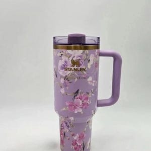 May include: A lavender Stanley tumbler with a floral design featuring pink and purple flowers. The tumbler has a handle and a lid. The text "STANLEY" and "Floral Fancy" are visible on the tumbler.