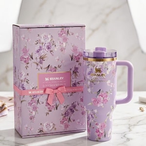 Puede incluir: Un vaso Stanley lavanda con asa y dise&ntilde;o floral, junto a una caja de regalo a juego. El vaso y la caja presentan flores rosas y moradas. La caja est&aacute; atada con una cinta rosa y tiene el texto "STANLEY LOVESHACKFANCY".