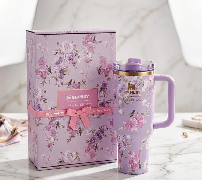 Puede incluir: Un vaso Stanley lavanda con asa y dise&ntilde;o floral, junto a una caja de regalo a juego. El vaso y la caja presentan flores rosas y moradas. La caja est&aacute; atada con una cinta rosa y tiene el texto "STANLEY LOVESHACKFANCY".
