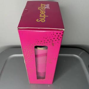 K&ouml;nnte beinhalten: Eine rosa Schachtel mit dem Wort "SuperStar" in goldener Schrift. Die Schachtel hat ein Fenster, das einen rosa Becher zeigt. Die Schachtel ist mit goldenen Sternen verziert.