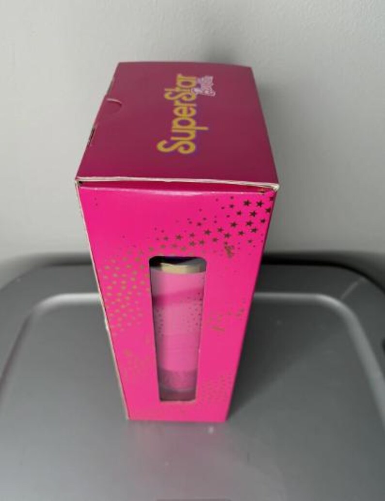 K&ouml;nnte beinhalten: Eine rosa Schachtel mit dem Wort "SuperStar" in goldener Schrift. Die Schachtel hat ein Fenster, das einen rosa Becher zeigt. Die Schachtel ist mit goldenen Sternen verziert.