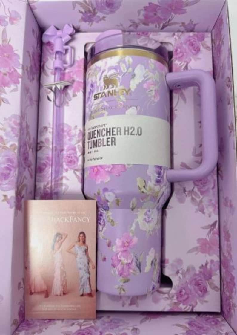 May include: A lavender Stanley Quencher H2.0 Tumbler with a floral design, a matching straw, and a book in a floral-patterned box. The tumbler has a handle and the text "QUENCHER H2.0 TUMBLER".