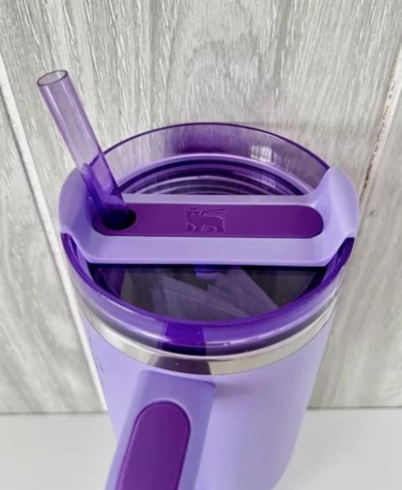 Op de afbeelding: Een lavendelkleurige ge&iuml;soleerde beker met een paars deksel en rietje. Het deksel heeft een schuifsluiting en een handvat. De beker heeft een roestvrijstalen band en een logo op het deksel.