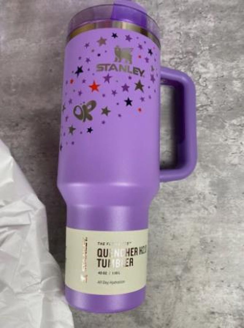 Op de afbeelding: Een paarse Stanley Quencher H2.0 tumbler met een handvat, versierd met sterren en een vlinder. De tumbler heeft een rietje en een capaciteit van 1,18L. De tekst "STANLEY" is zichtbaar.