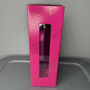 K&ouml;nnte beinhalten: Eine leuchtend pinke Schachtel mit goldenen Sternen und einem rechteckigen Fenster, das ein lila Produkt zeigt. Das Produkt scheint ein Beauty-Artikel zu sein, m&ouml;glicherweise ein Parf&uuml;mflakon oder ein &auml;hnliches Kosmetikprodukt.