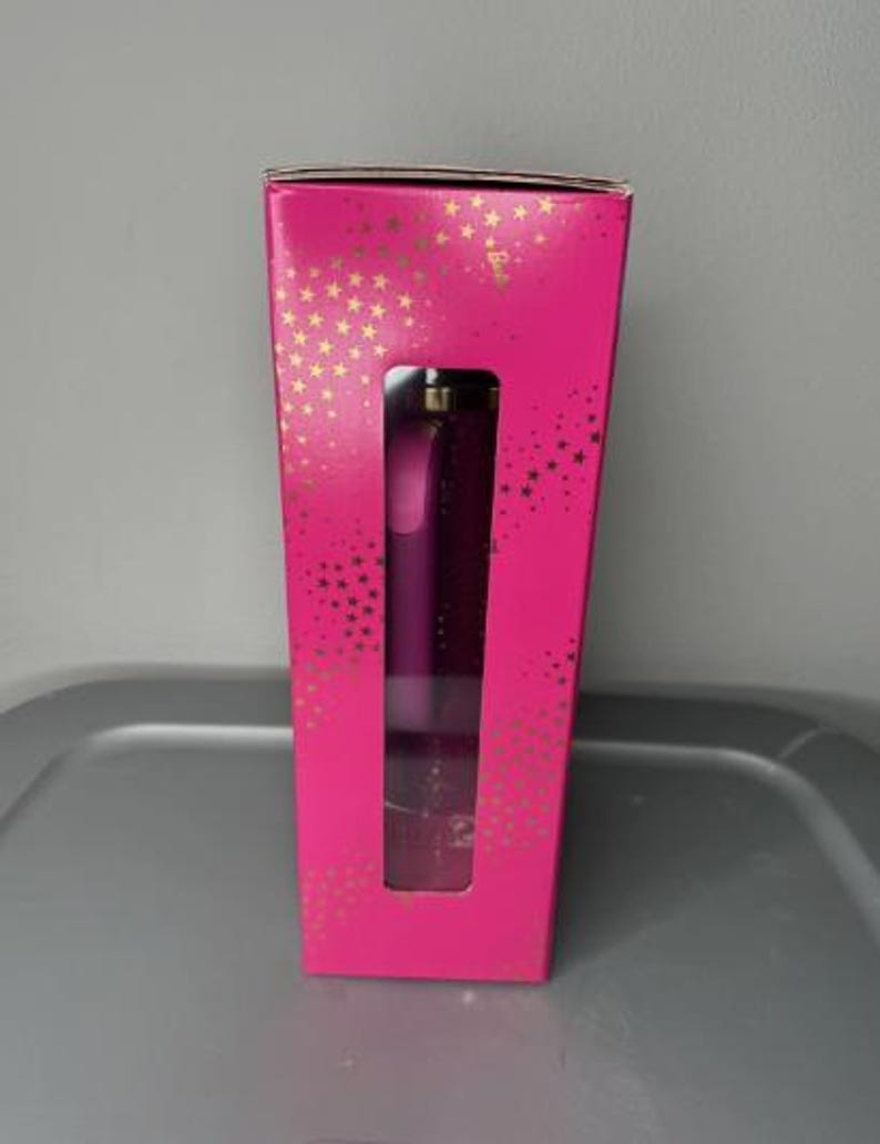 K&ouml;nnte beinhalten: Eine leuchtend pinke Schachtel mit goldenen Sternen und einem rechteckigen Fenster, das ein lila Produkt zeigt. Das Produkt scheint ein Beauty-Artikel zu sein, m&ouml;glicherweise ein Parf&uuml;mflakon oder ein &auml;hnliches Kosmetikprodukt.