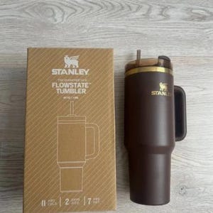 May include: A brown Stanley Flowstate tumbler with a handle and straw, alongside its cardboard packaging. The box displays the Stanley logo and product information. The tumbler is designed to keep drinks cold for 2 days and hot for 7 hours.