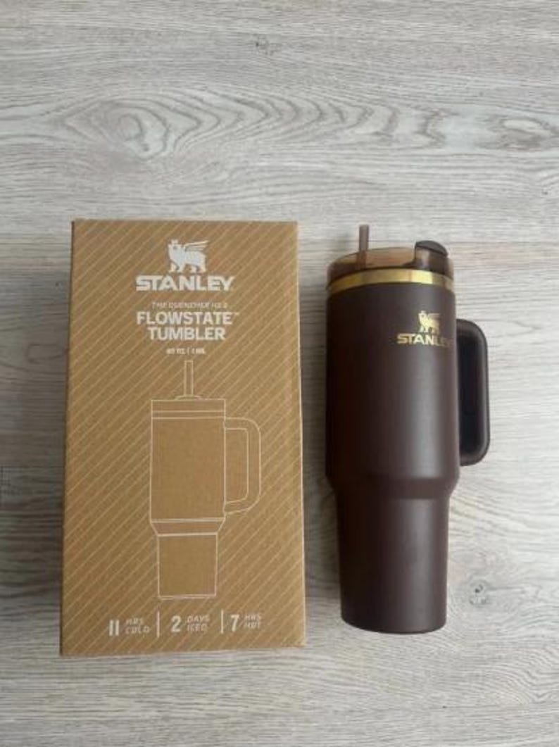 May include: A brown Stanley Flowstate tumbler with a handle and straw, alongside its cardboard packaging. The box displays the Stanley logo and product information. The tumbler is designed to keep drinks cold for 2 days and hot for 7 hours.