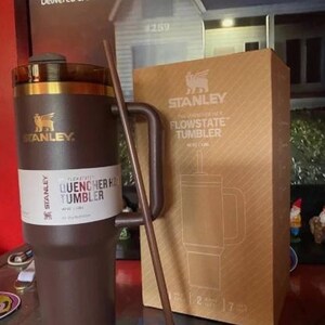 May include: A brown Stanley Quencher H2.0 Flowstate tumbler with a matching straw and a cardboard box. The tumbler has a handle and a white label with product information. The box features the Stanley logo and a diagram of the tumbler.
