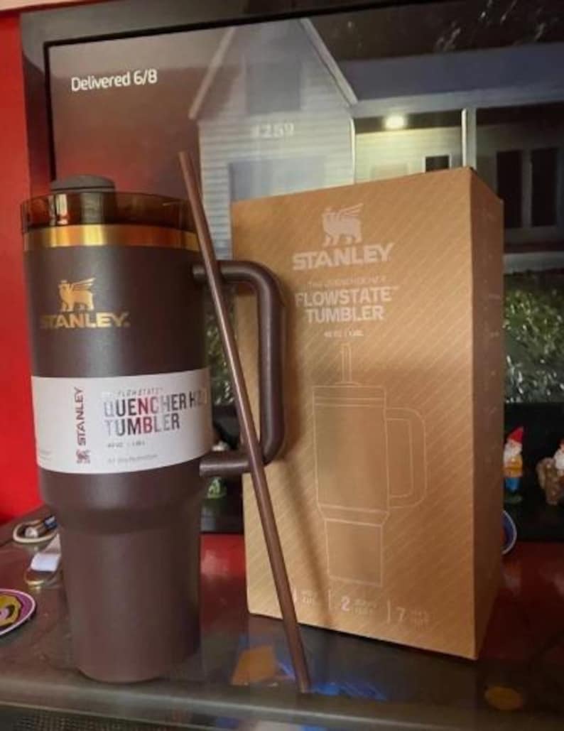 May include: A brown Stanley Quencher H2.0 Flowstate tumbler with a matching straw and a cardboard box. The tumbler has a handle and a white label with product information. The box features the Stanley logo and a diagram of the tumbler.