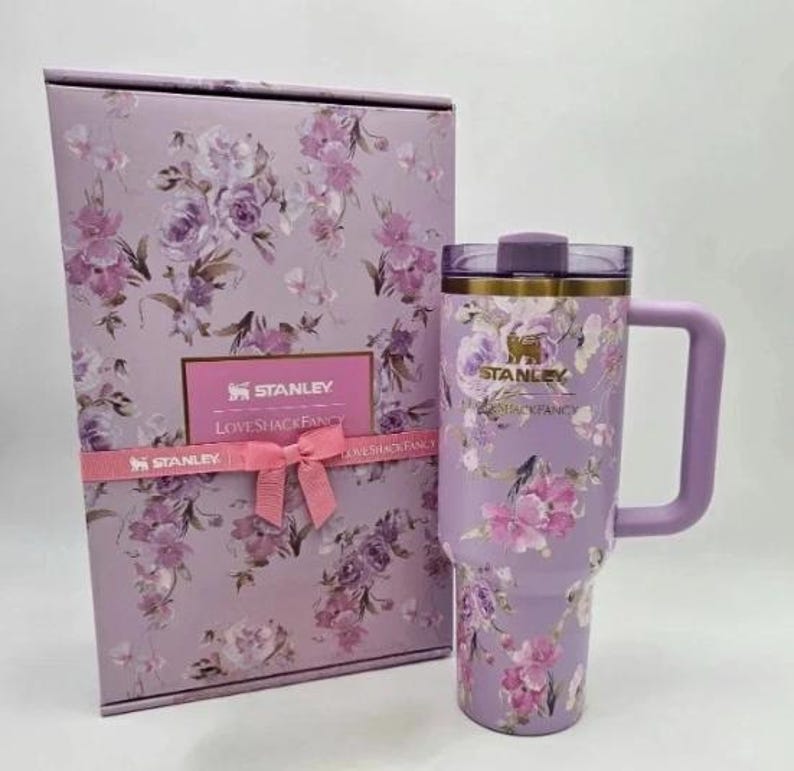 May include: A lavender Stanley tumbler with a handle and a matching gift box. Both the tumbler and box feature a floral design with pink and purple flowers. The box has a pink ribbon and the Stanley and LoveShackFancy logos.