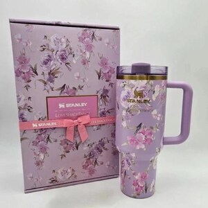Puede incluir: Un vaso Stanley lavanda con asa y caja de regalo a juego. Tanto el vaso como la caja presentan un dise&ntilde;o floral con flores rosas y moradas. La caja tiene una cinta rosa con el logo de Stanley.