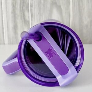 Op de afbeelding: Een paarse beker met bijpassend deksel en rietje. Het deksel heeft een lichtpaarse handgreep en een donkerpaarse band met een gevleugeld paardenlogo. Ontworpen voor koude dranken.