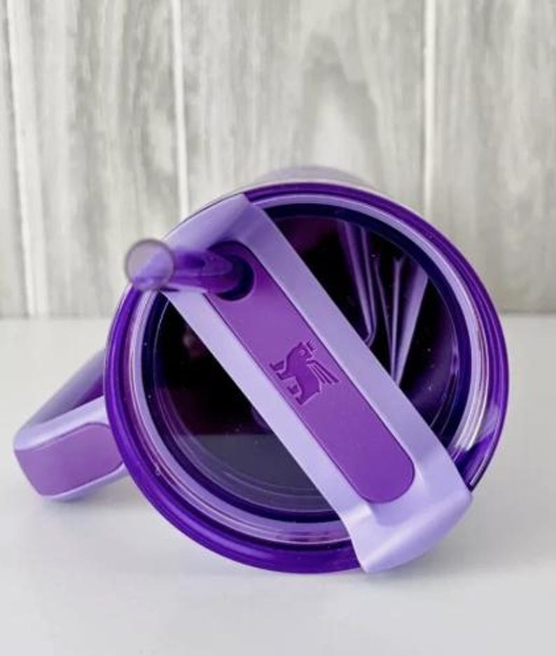 Op de afbeelding: Een paarse beker met bijpassend deksel en rietje. Het deksel heeft een lichtpaarse handgreep en een donkerpaarse band met een gevleugeld paardenlogo. Ontworpen voor koude dranken.