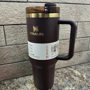 May include: A dark brown Stanley Quencher tumbler with a handle. The tumbler has a gold-coloured rim and a black lid. The Stanley logo is visible in gold. A white label with text is wrapped around the tumbler.