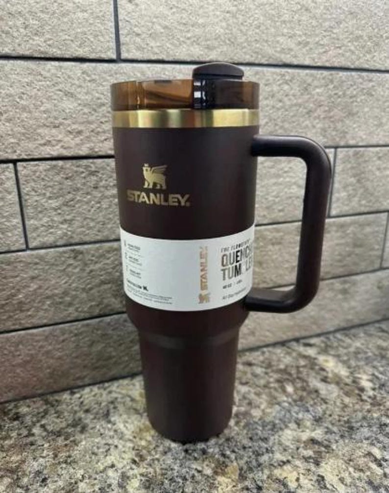 May include: A dark brown Stanley Quencher tumbler with a handle. The tumbler has a gold-coloured rim and a black lid. The Stanley logo is visible in gold. A white label with text is wrapped around the tumbler.
